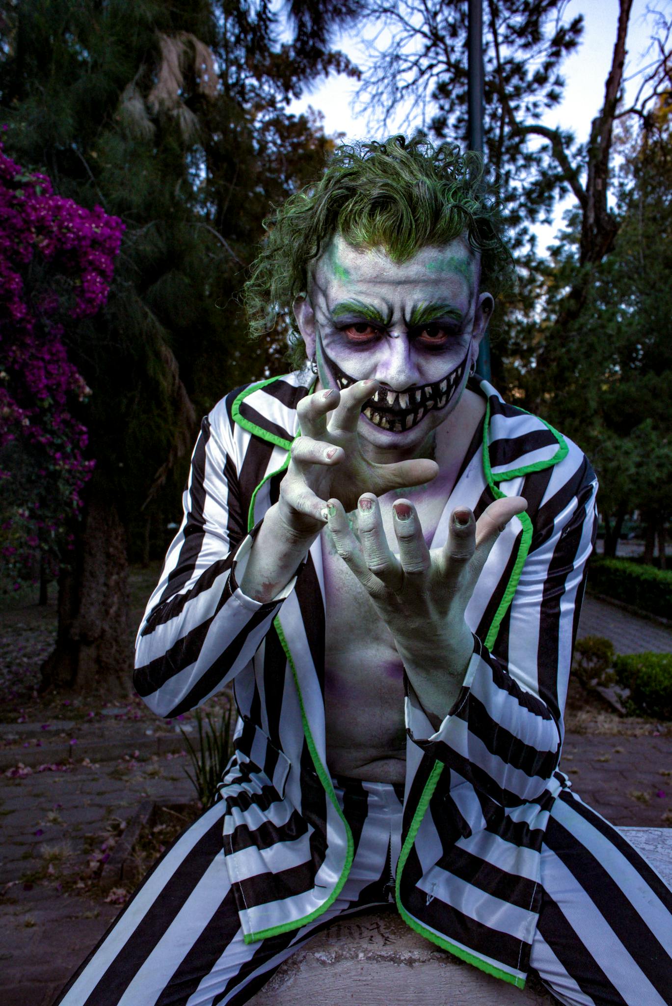 Creepy clown in green makeup and striped suit posing menacingly in a forest setting.